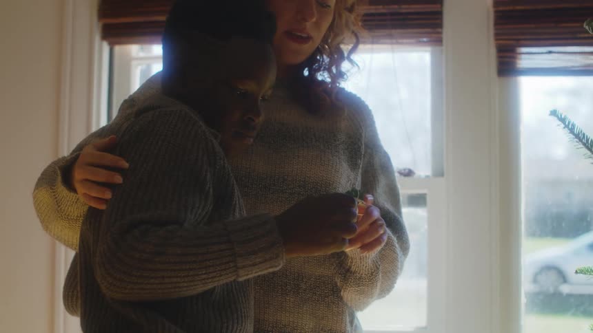Mother hugs child while showing them christmas ornament as they stand near christmas tree