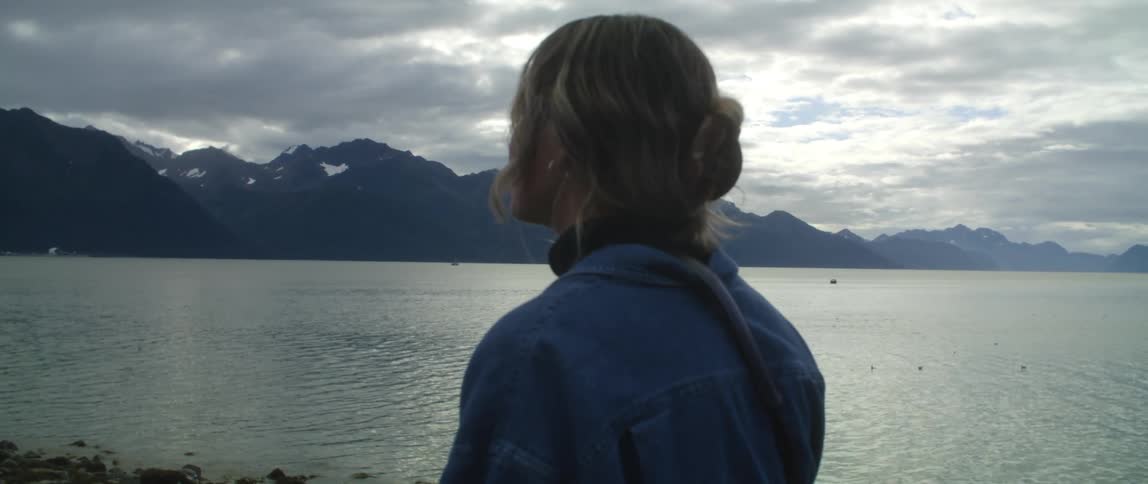 A woman looking out over a lake from the rocky shore turns her head to look into the camera