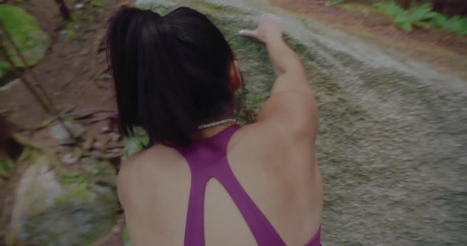 Woman with ponytail and purple sports bra climbs up large rock in the forest