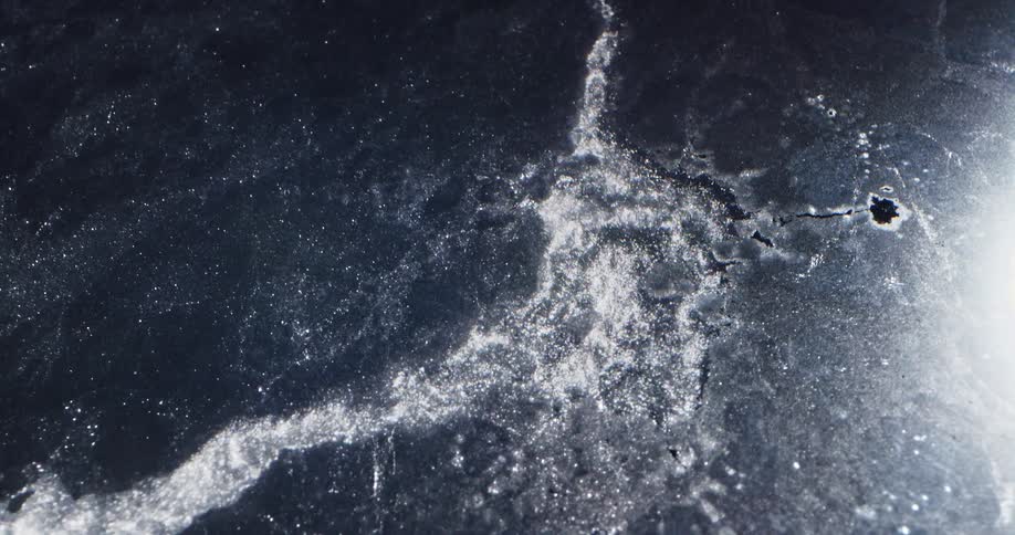 Water flows through gray shiny ice sheet pulling pieces off of it