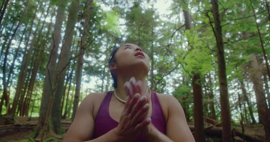 A woman is standing in the woods and looking up at the trees as she rubs her hands together