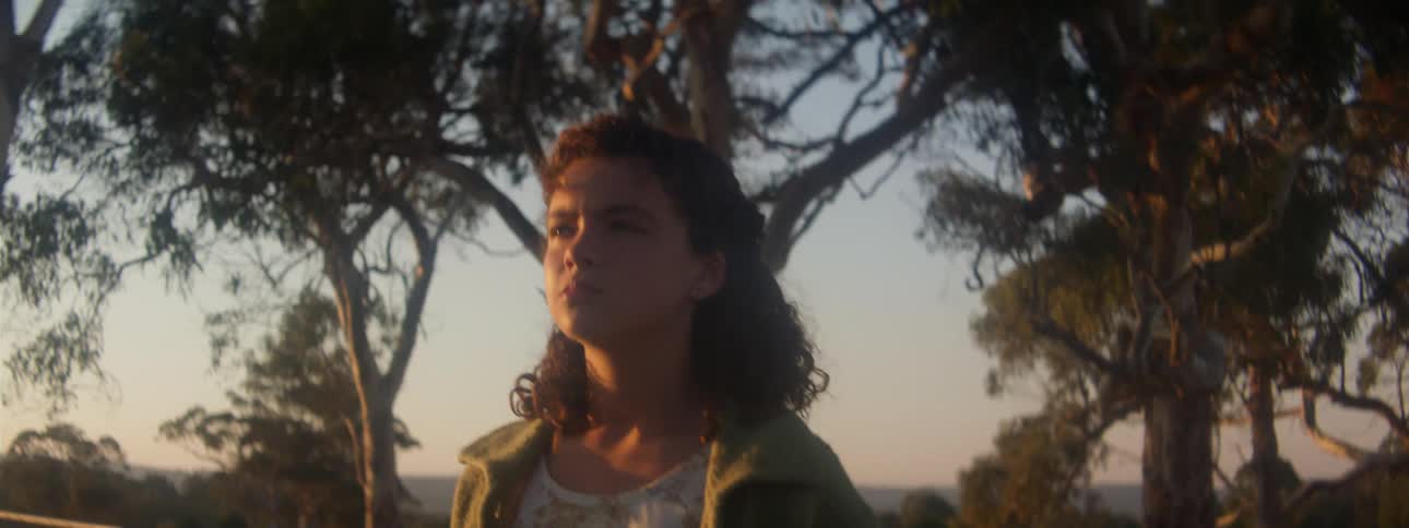 Woman with curly hair green jacket and white blouse walks in the field at sunset