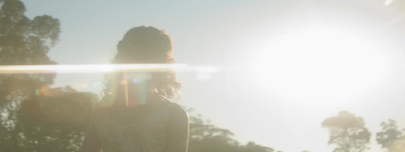 The sun is shining brightly upon a girl standing outside looking at trees under a clear sky
