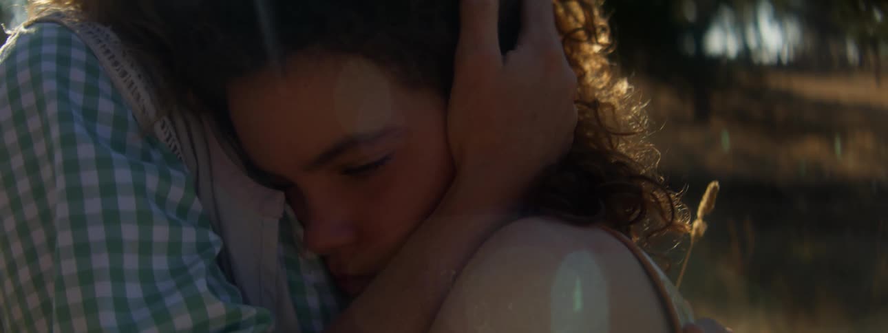 Woman with brown hair and plaid shirt holds woman with curly hair in the field