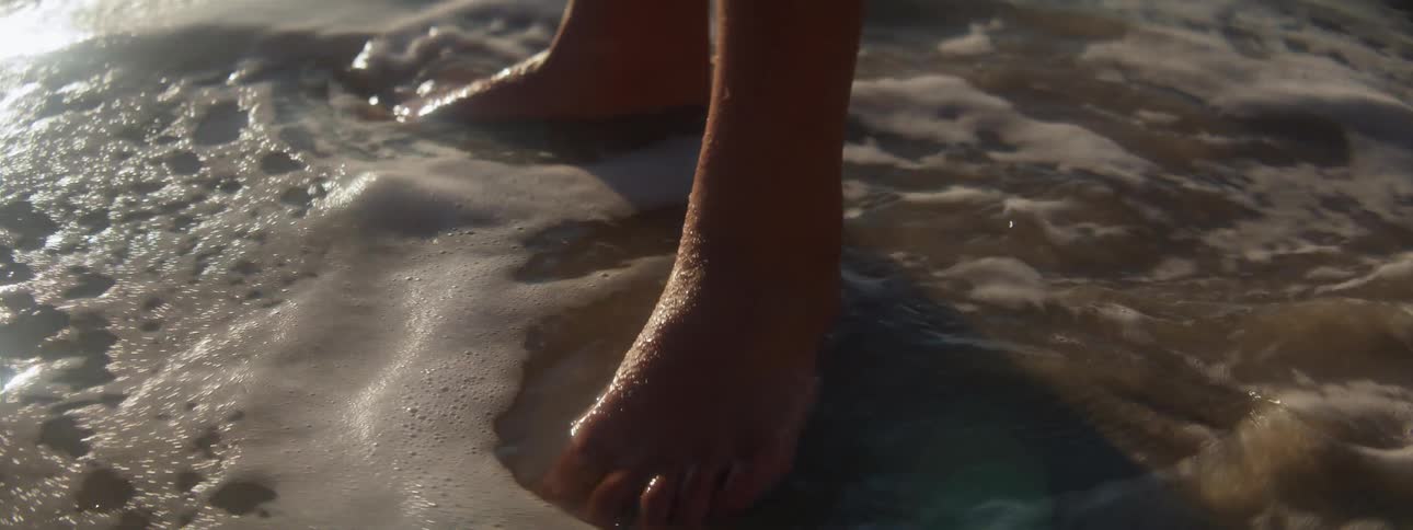 Ocean waves washing around a persons bare feet on a sandy beach