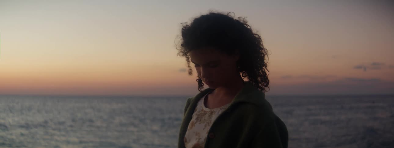 A girl is standing outside near the water as waves roll in and the wind gently blows her hair