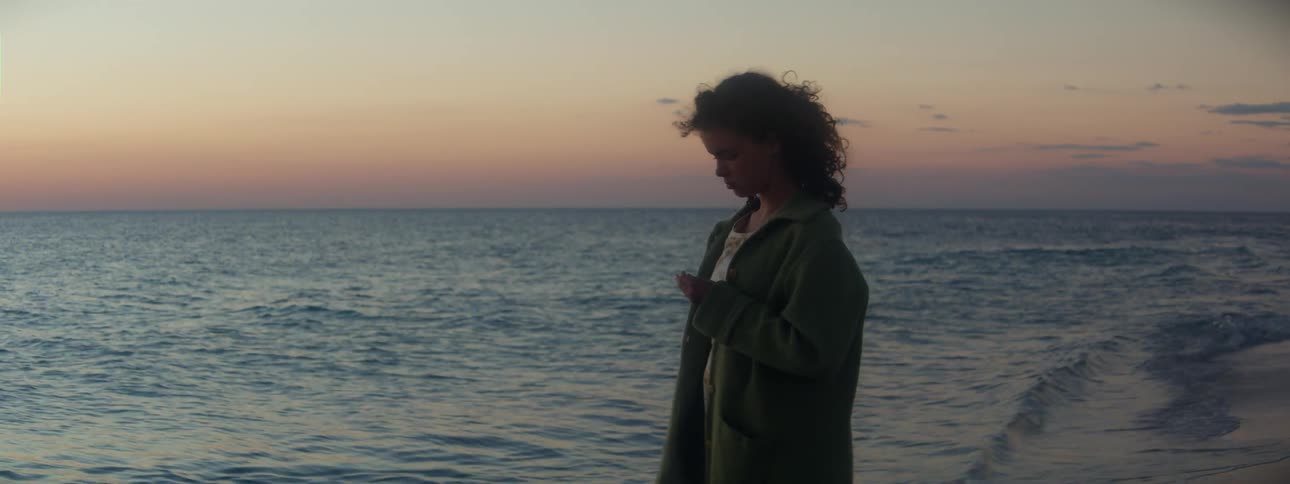 A girl is standing outside near the water as waves roll in and the wind gently blows her hair