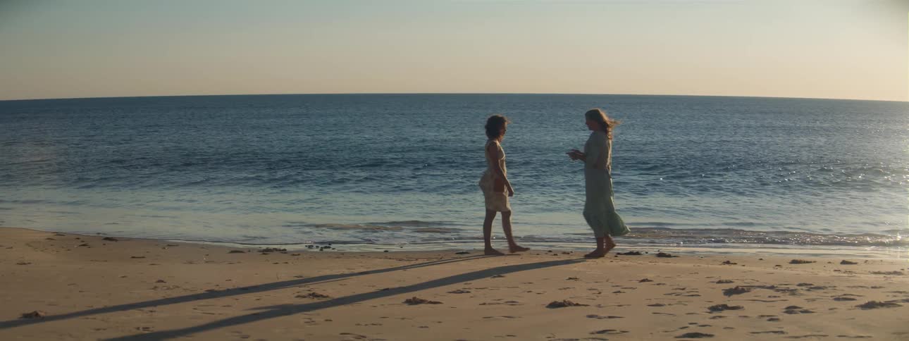 A woman in a sundress picks up a rock on the beach and walks over to her teenage daughter and puts it in her hands as the waves roll into shore