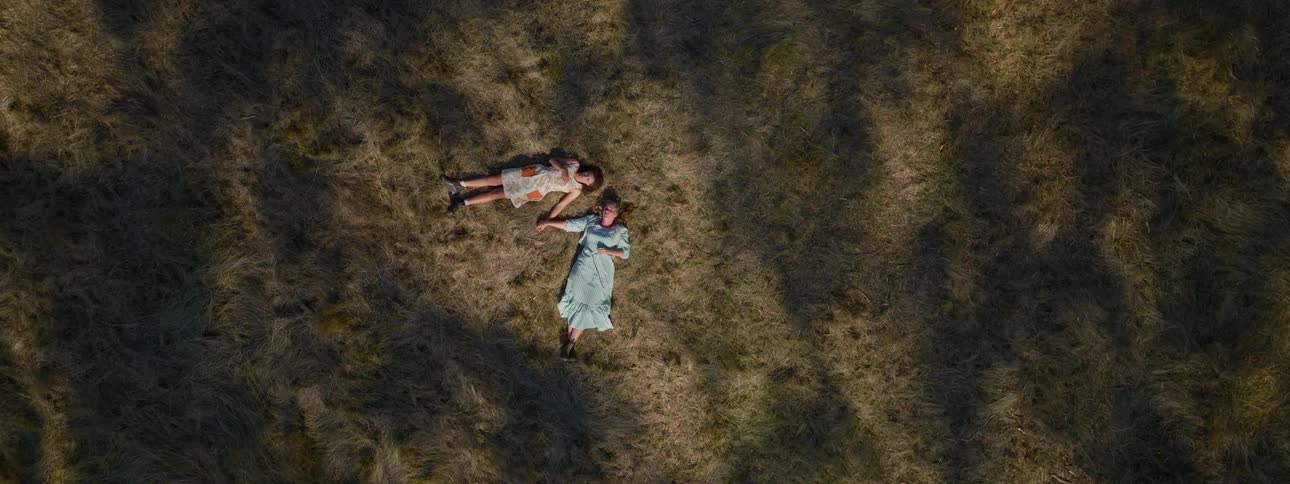 Women in light blue and white dresses lie down on brown grass holding hands in the field