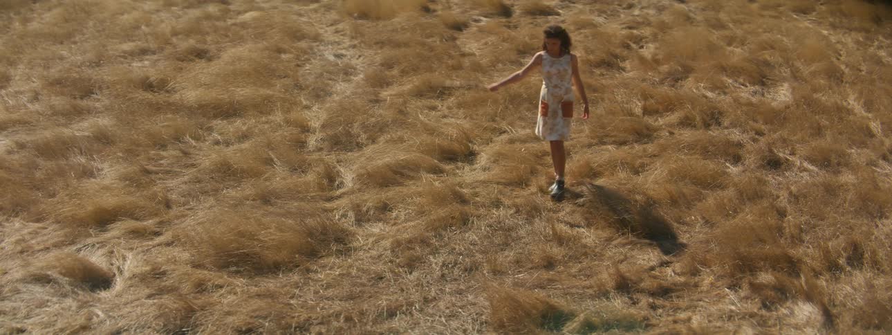 Woman with curly hair white dress and a pair of boots walks in the field swaying a stick