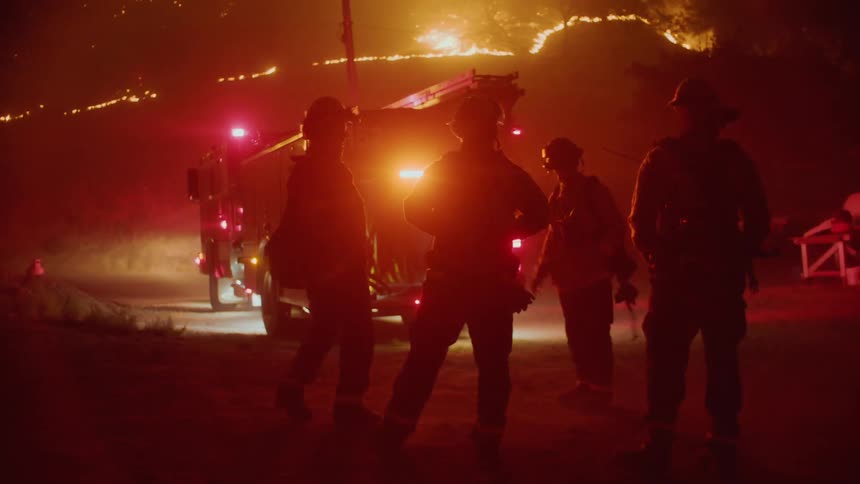 A group of firefighters in bunker gears stands talk with each other near the firetruck at night