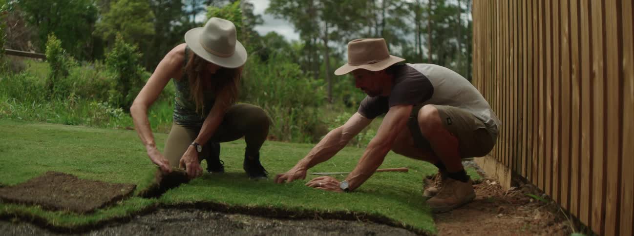 A man and woman are both wearing sunhats as they kneel in their yard and lay down sod