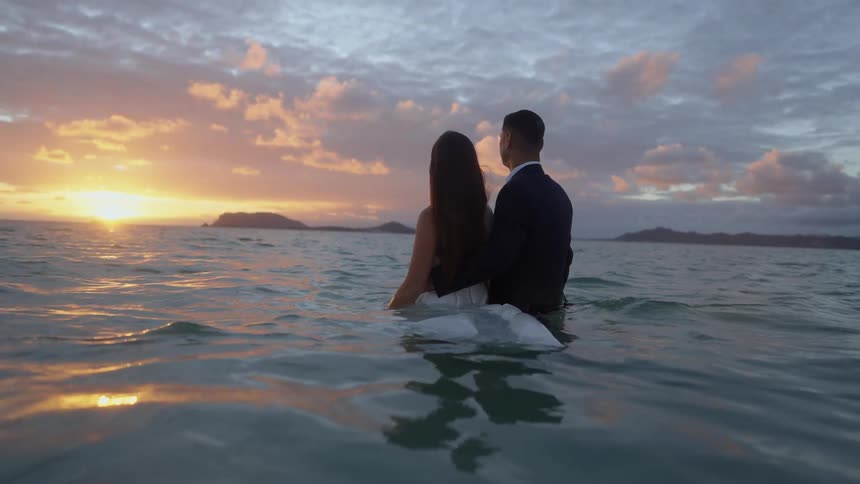 Bride in wedding dress and groom in dark suit stand in ocean water watching sunset