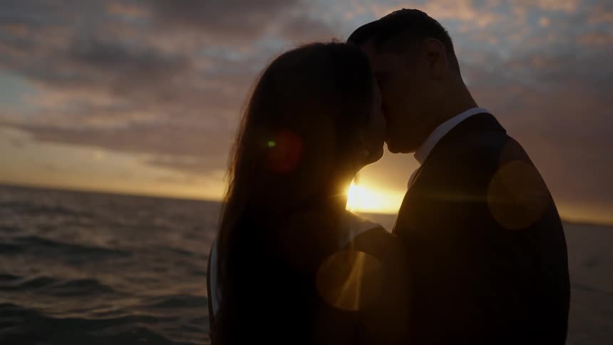 Silhouette of a man and woman standing near the water kissing as the setting sun reflects on the ripples in the water