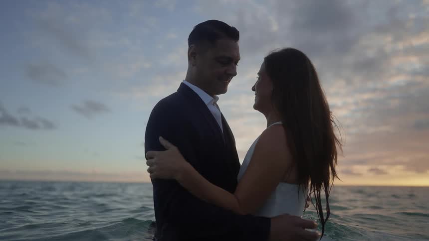 A man and a woman are smiling with their arms around each other as they stand in the water at the beach