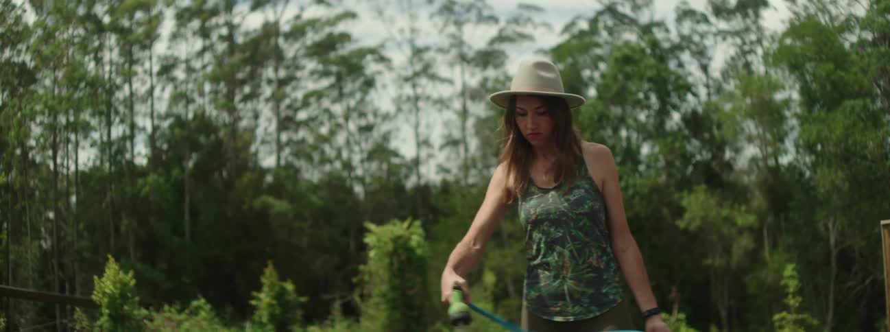 A woman is wearing a sunhat as she walks around in her yard holding a hose as she waters her plants