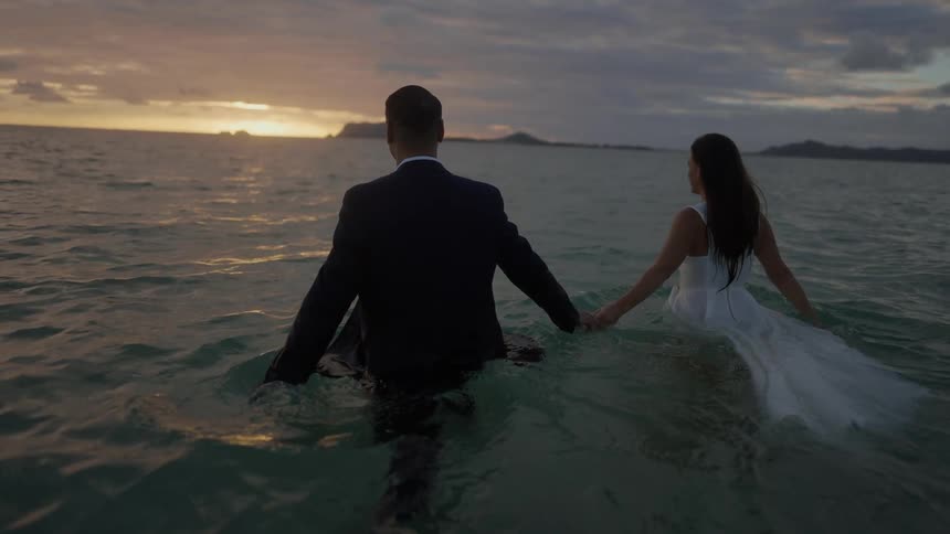Groom in black suit and bride wearing wedding dress walk in sea at sunset
