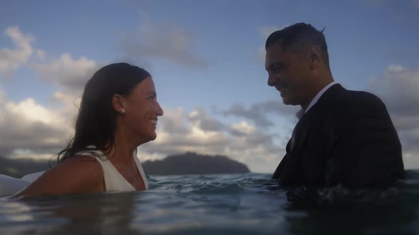 A man and woman fully clothed stand in the ocean together and smile at one another