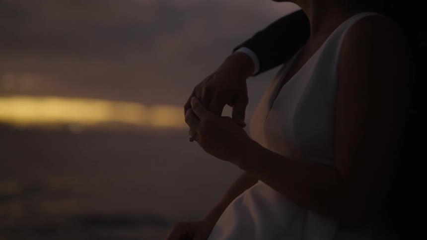 Groom in black suit puts arm on brides shoulder travelling on boat at sunset