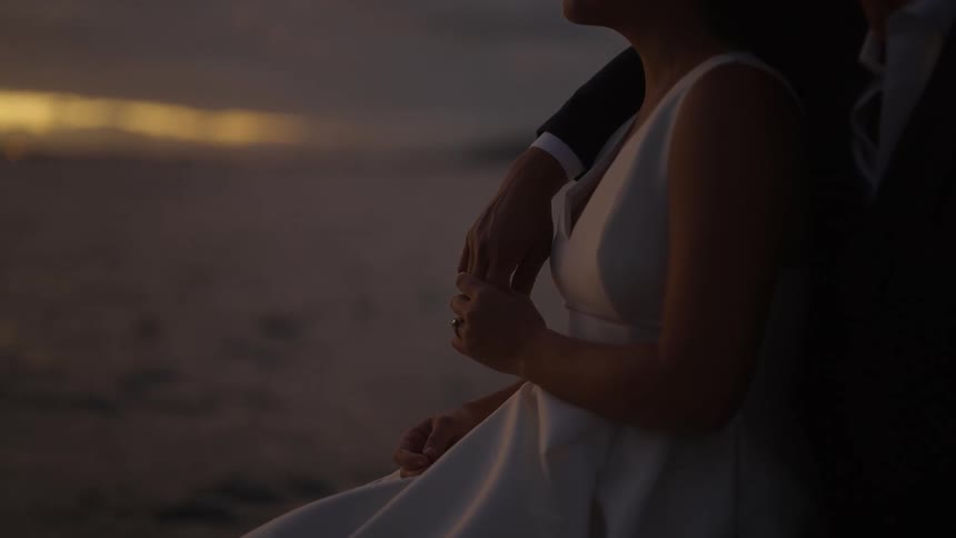 A bride and groom hold hands in front of the ocean as the sun sets