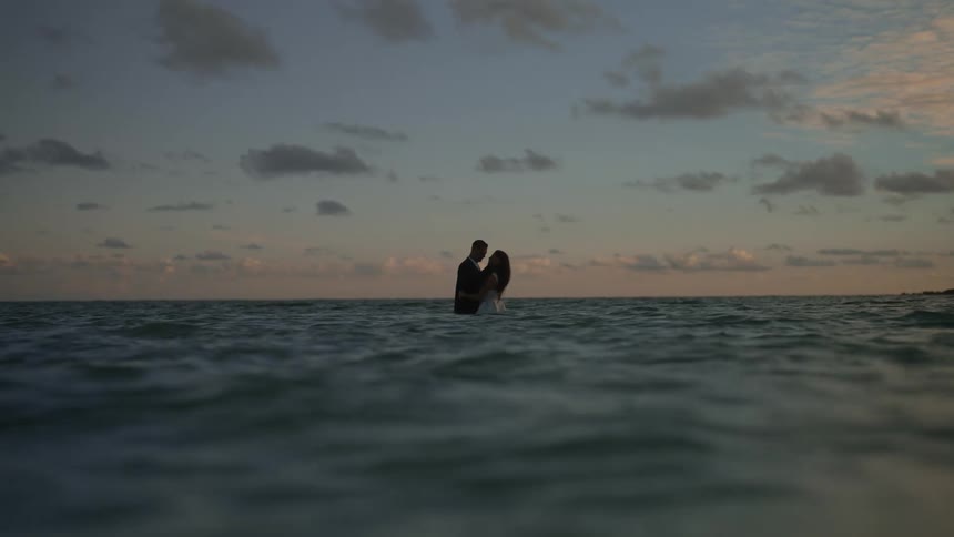 A man and a woman are standing in the water at the beach with their arms around each other and they kiss