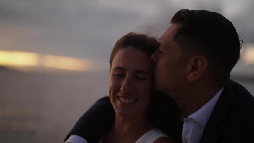 A man and a woman are standing near the water as waves roll in and she smiles as he kisses her forehead