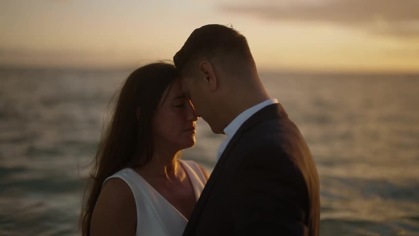 A man and woman stand on the shore with their foreheads resting against one another