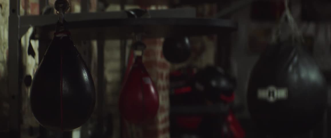 Many punching bags are hanging in a boxing gym as the lights go off