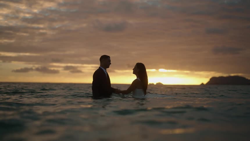 A man and woman in formal clothes stand in the ocean and walk towards one another and take hands and kiss
