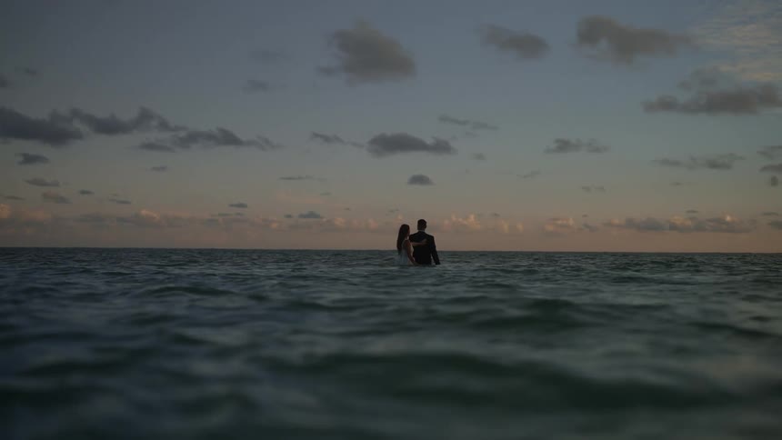 Bride in wedding dress and groom in suit walk in ocean water at sunset