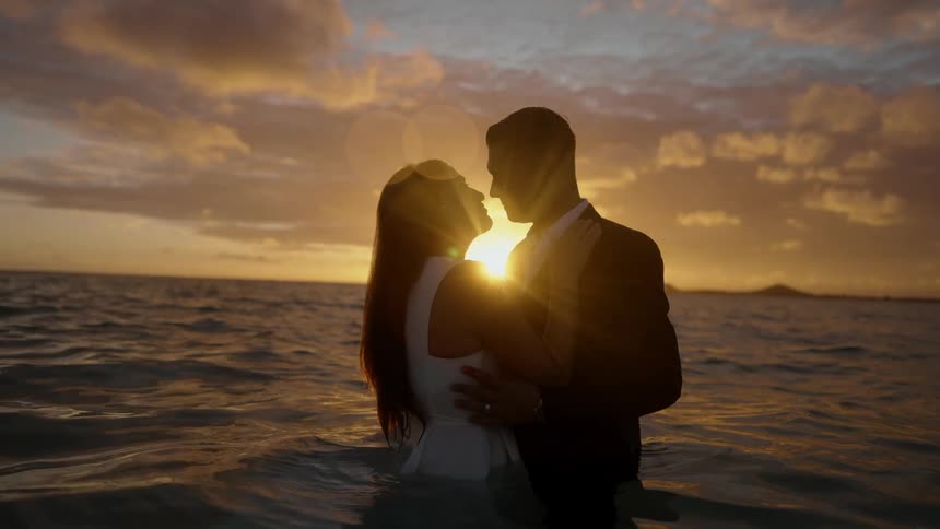 A man and woman fully clothed stand in the ocean and hold one another as they look in to each others eyes