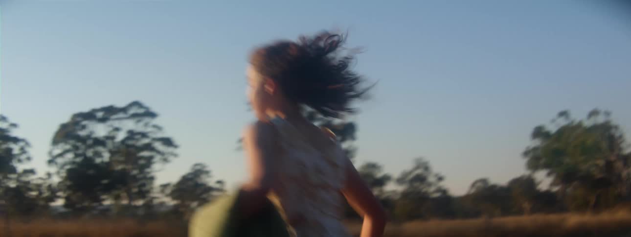 The sun is shining brightly upon a girl running outside with trees in the distance under a clear sky