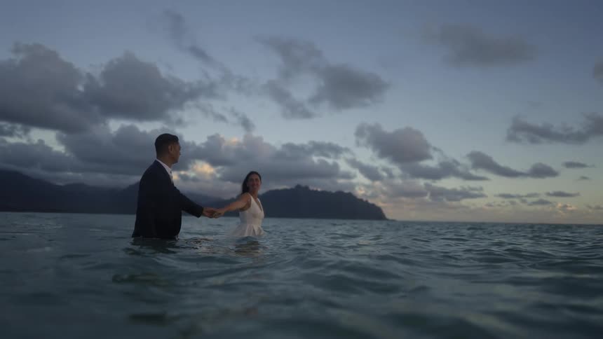 A bride and groom hold hands as they walk in the ocean