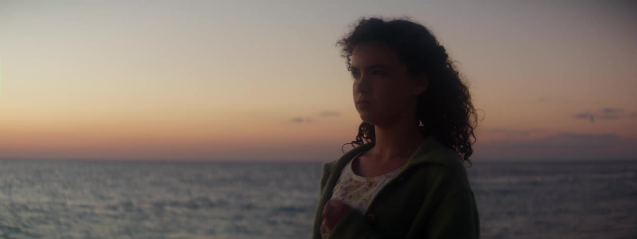 A girl is standing outside near the water as waves roll in and the wind gently blows her hair