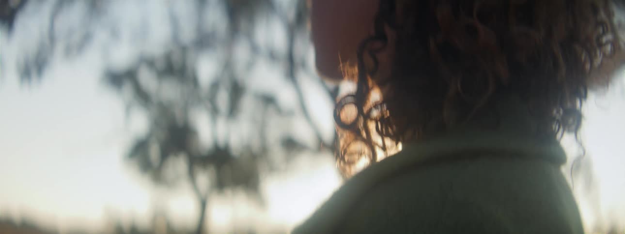 Woman with curly hair and green jacket walks in the field looking around at sunset