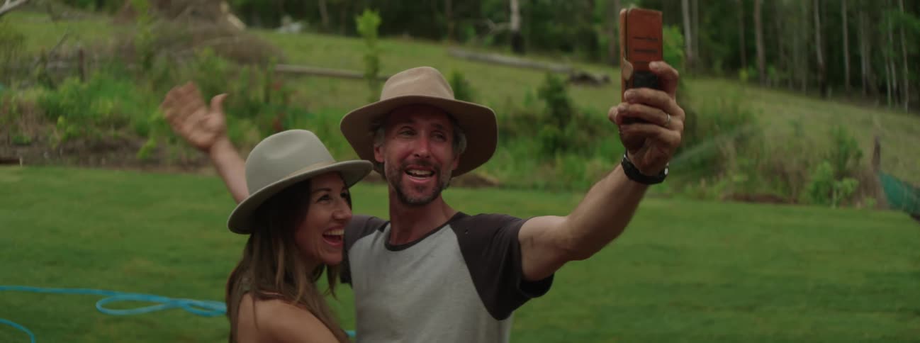 Man with hat beard and woman with hat and brown hair take selfie with smartphone in green backyard kissing and smiling