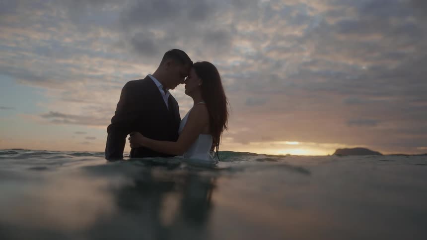 A man and a woman are staring into each others eyes and she has her arms around him as they stand in the water at the beach