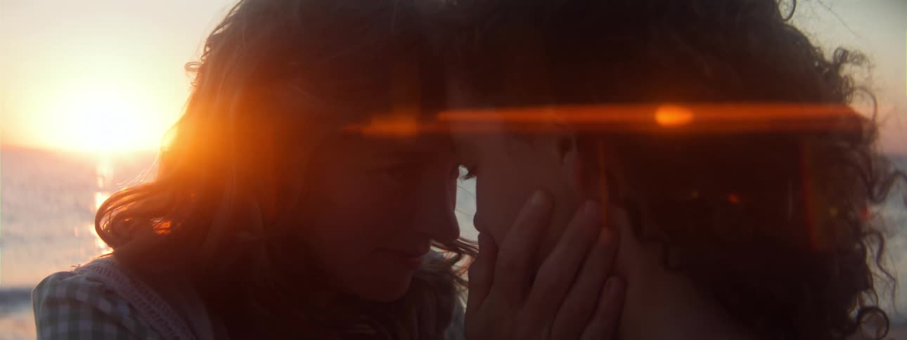 Blonde woman and woman with brown wavy hair hold each other and look into each others eyes on the beach near the ocean