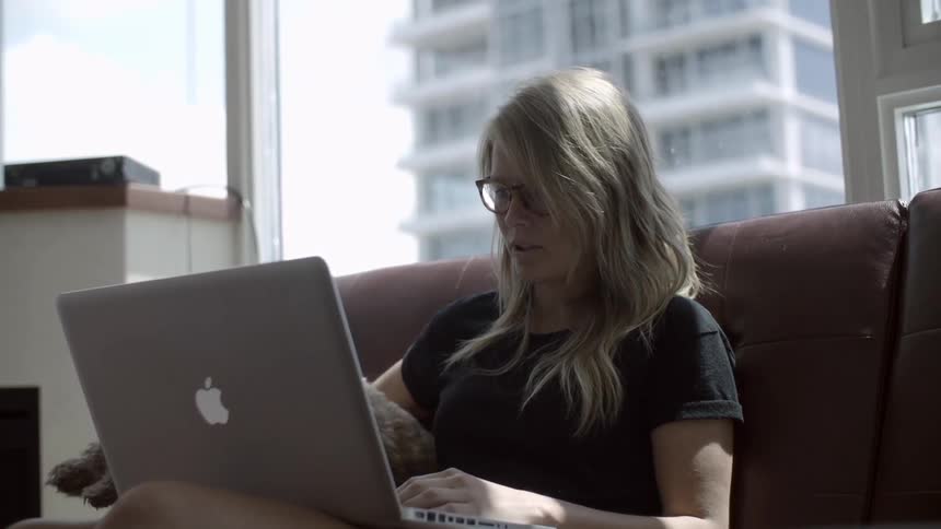 A woman sits on her sofa with her laptop and her dogs jumps up to sit with her