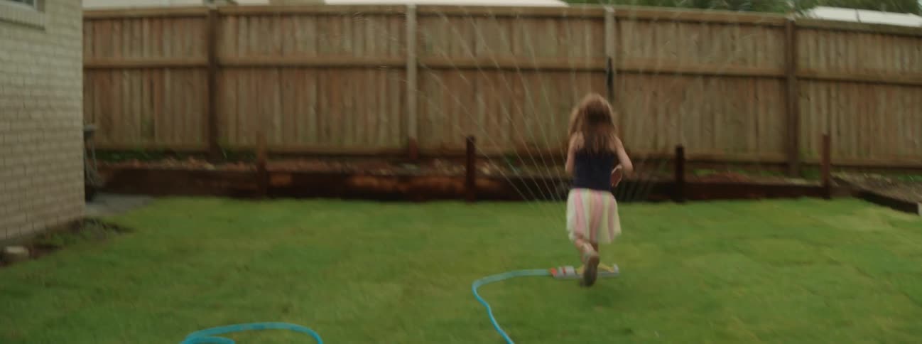 A young girl with red hair in a tutu runs through a sprinkler in her backyard
