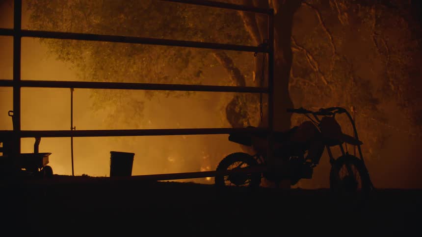 A silhouette of a motorcycle against a fence with smoke and flames in the background