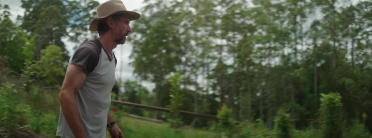 A gardener wearing a hat walking a wheelbarrow through a yard