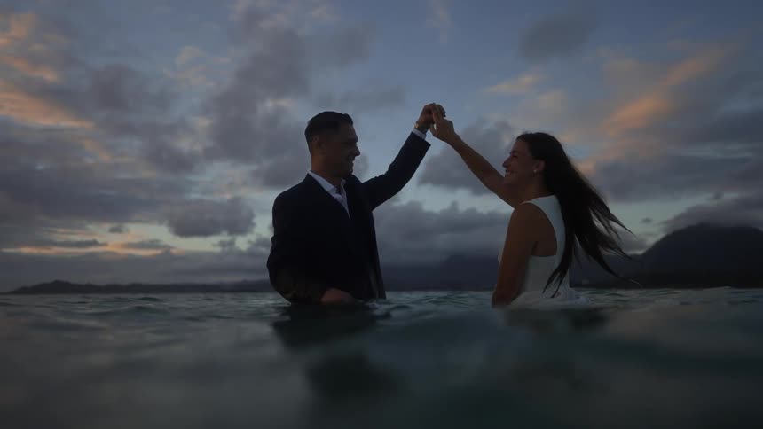 A married couple in formal attire dancing waist deep in water outside