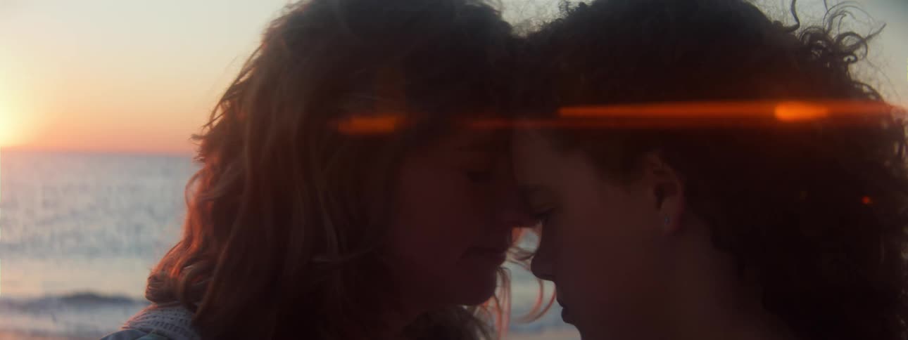 Blonde woman kisses womans forehead on the beach at sunset next to the sea