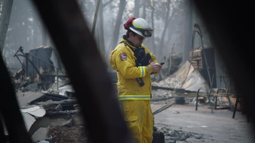 A firefighter in a yellow suit and white helmet stands texting among rubble and debris