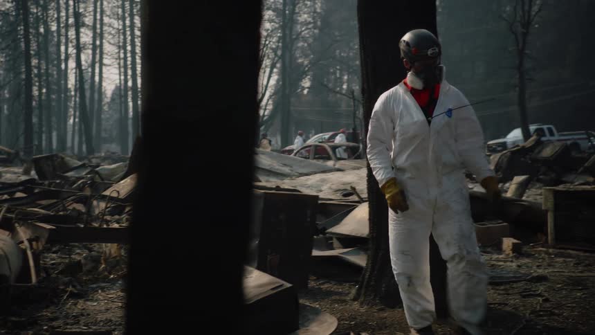 An emergency response team in white jumpsuits are investigating the debris where a building once stood as smoke lingers in the air