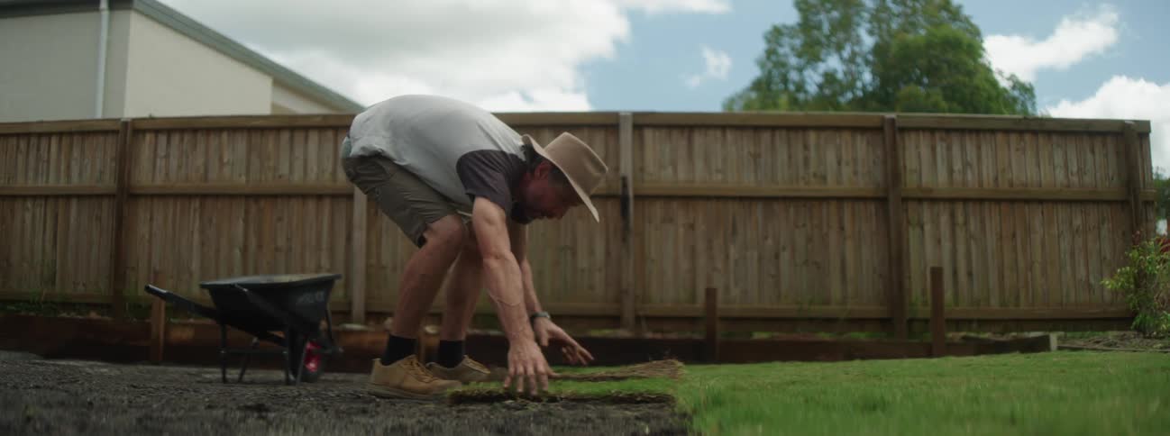 A gardener in a hat lays out a piece of grass against the rest of the yard