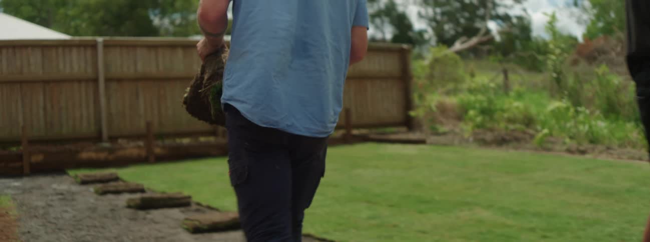 A man in a blue shirt grabs a layer of sod grass and drops it down on the ground