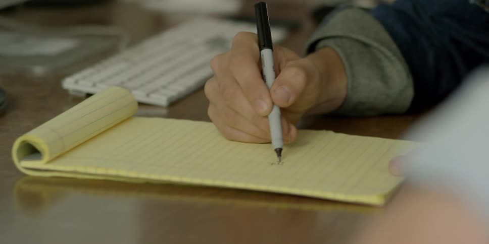 Two people sit at a desk while one sketches something out on a yellow pad using a pen