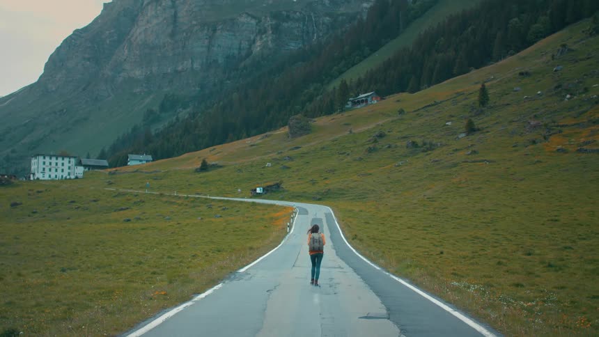 A woman wearing a backpack walks down a road in a mountain valley during the day